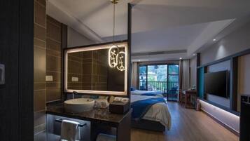 A modern hotel room with a large mirror, a round sink, a bed with a blue blanket, a TV mounted on the wall, and a balcony with a view.