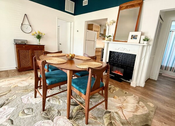Un coin repas avec une table en bois, des chaises rembourrées bleues, un tapis décoratif, une cheminée et, au fond, une cuisine.