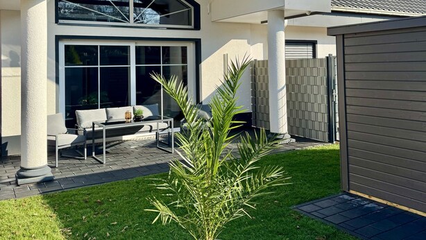 A modern house with a large glass window, a patio with outdoor furniture, and a potted plant in the foreground.
