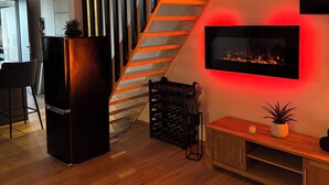 A modern living room with a wooden staircase, a black refrigerator, a TV mounted on the wall, and a wooden cabinet.