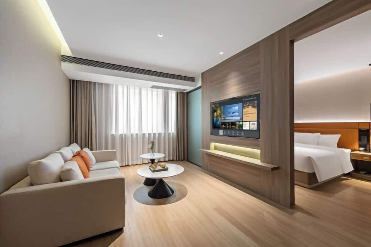 Una habitación de hotel moderna con sofá, una mesita redonda pequeña, televisor montado en un panel de madera y una cama con cabecero.