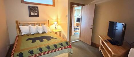Une chambre à coucher comprenant un lit, un téléviseur, une table de chevet et une porte donnant sur une autre pièce.