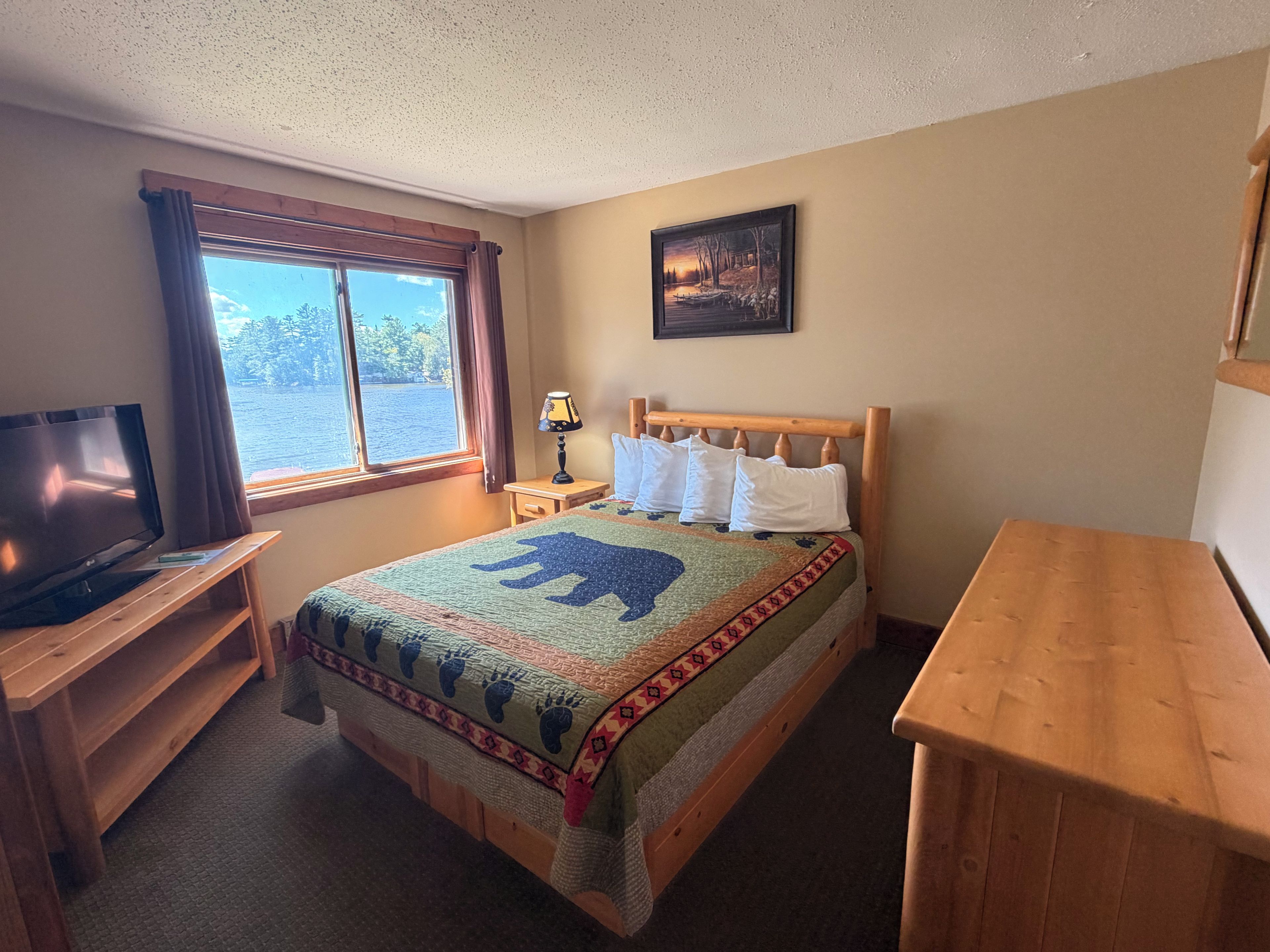 A room with a bed, a television on a wooden stand, a window with curtains, a lamp, and a painting on the wall.