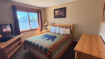 A room with a bed, a television on a wooden stand, a window with curtains, a lamp, and a painting on the wall.