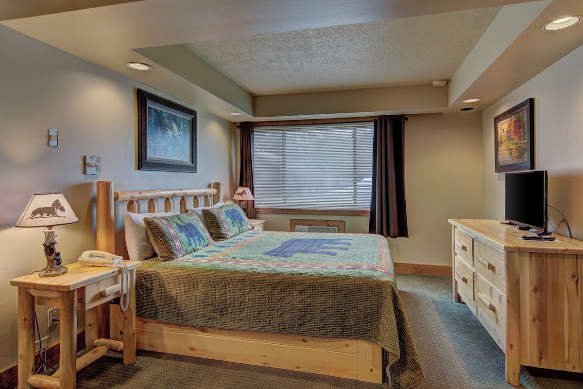 Un dormitorio con una cama de madera, una mesita de noche con lámpara, un aparador, un televisor y una ventana con persianas.