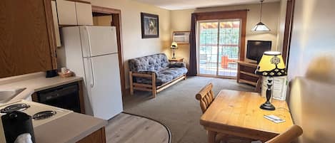 A compact kitchen with a refrigerator, stove, and wooden cabinets. A dining area with a table and chairs is adjacent to a sofa.