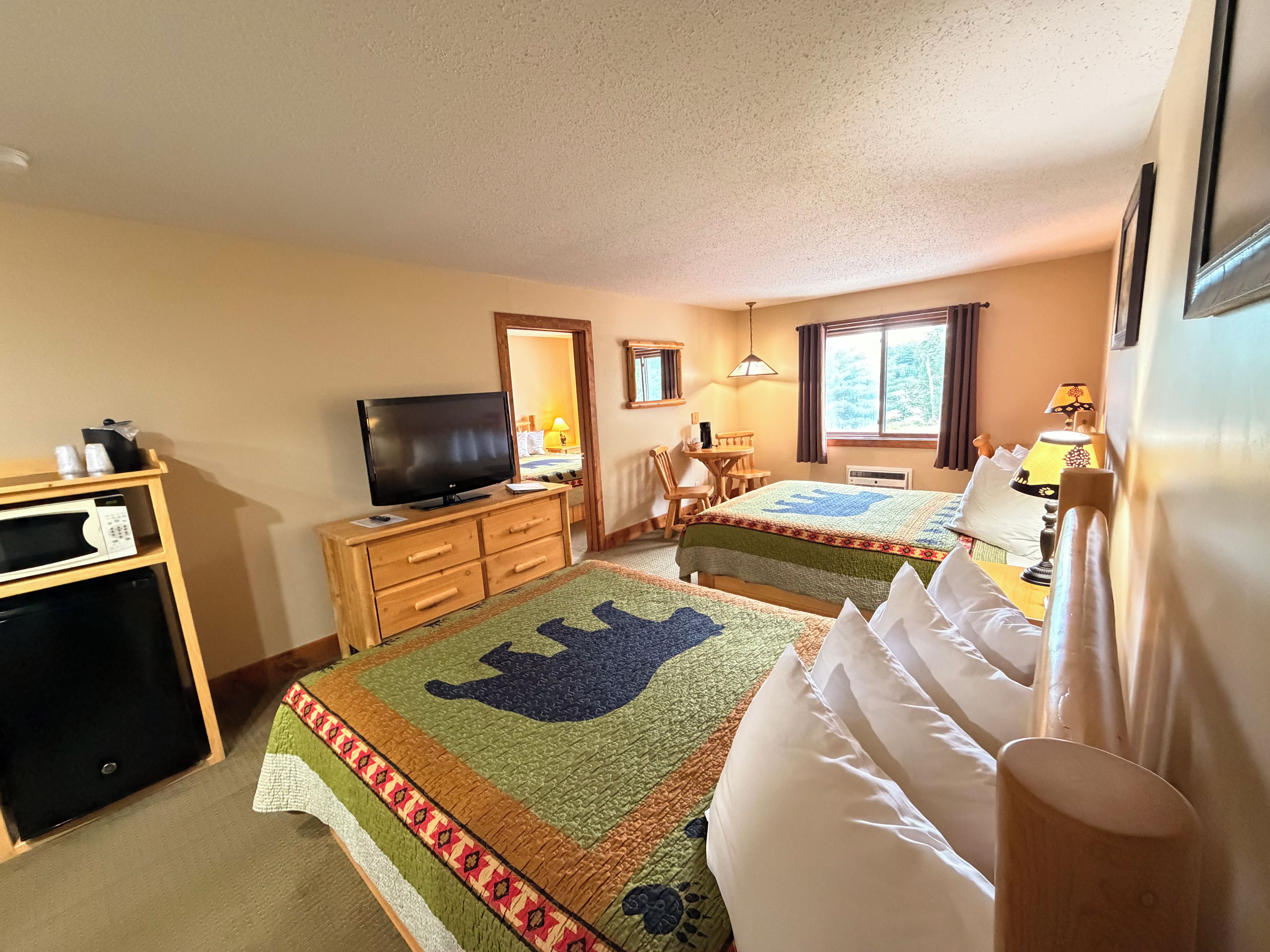 A hotel room with a television, a desk, a chair, two beds with quilts, a window, and a mirror.