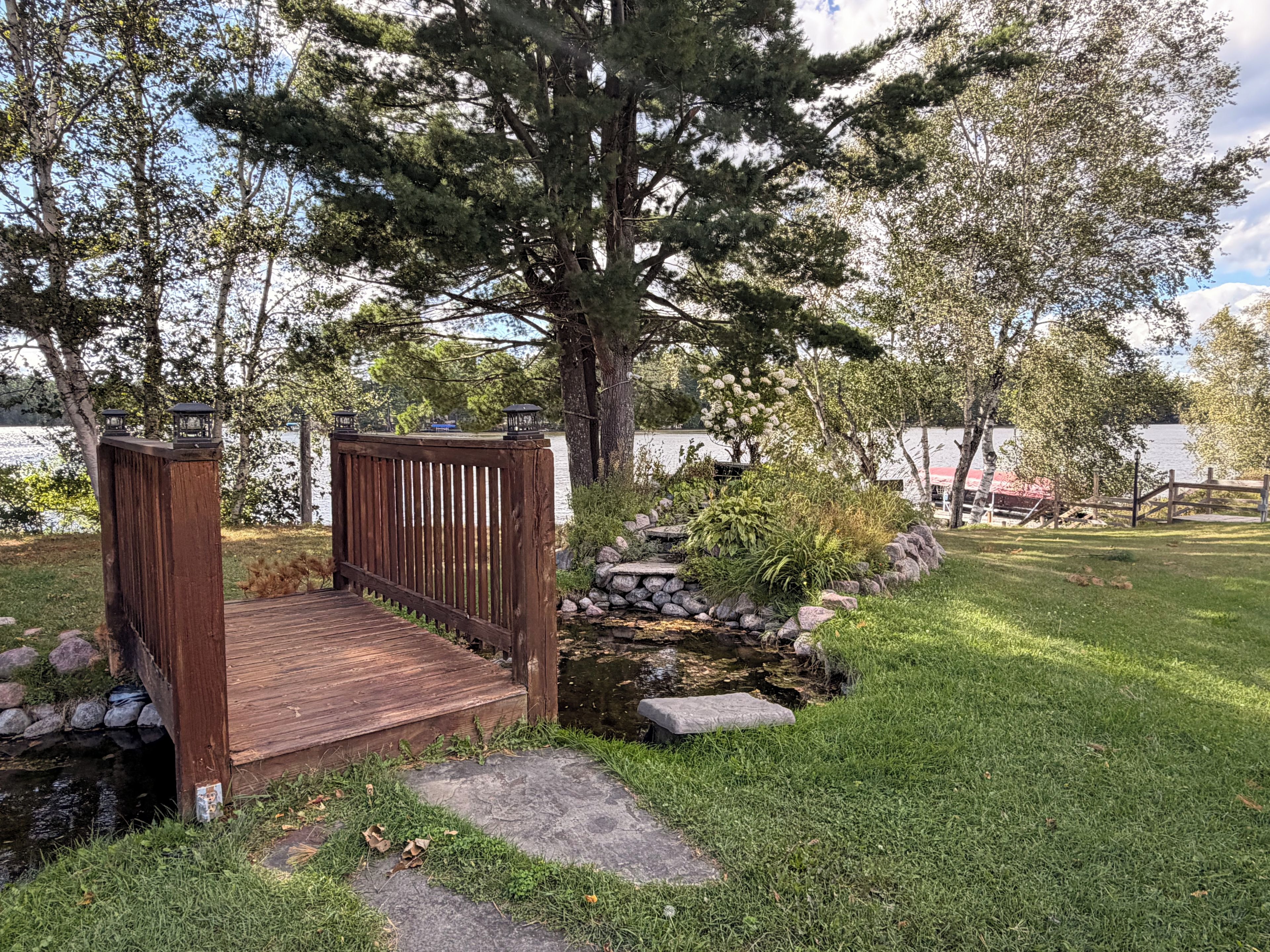 A wooden bridge leading to a dock over a small stream, surrounded by lush greenery and trees.