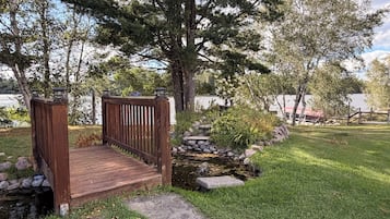 A wooden bridge leading to a dock over a small stream, surrounded by lush greenery and trees.