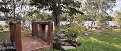 A wooden bridge leading to a dock over a small stream, surrounded by lush greenery and trees.