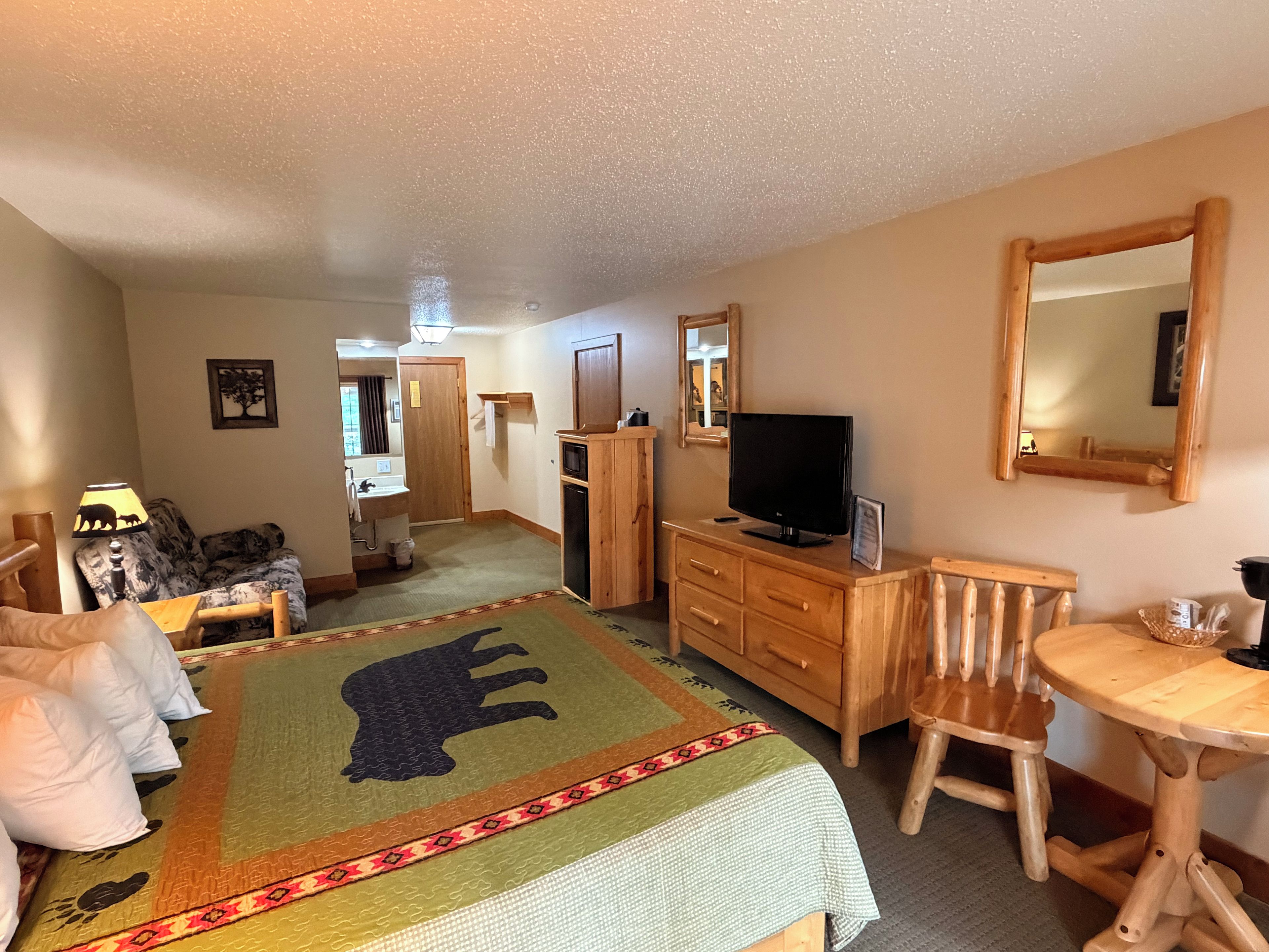 Una habitación de hotel con cama, televisión, cómoda de madera, silla, mesita y espejo.