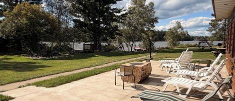 A patio with lounge chairs and a table, overlooking a lake with trees and a playground.