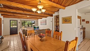 Un coin repas avec une table en bois, des chaises et un saladier rempli de fruits. Il y a également un espace cuisine équipé d’appareils électroménagers et doté d’une fenêtre.