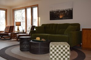 A living room with a green sofa, a checkered ottoman, a round coffee table with a candle, and a framed photograph on the wall.