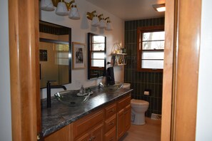 A bathroom with a glass bowl sink, wooden cabinets, a toilet, and a window with a view of trees.