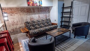 A living room with a patterned sofa, a coffee table, a black armchair, a red chair, a brick wall, and a door with a black ladder.