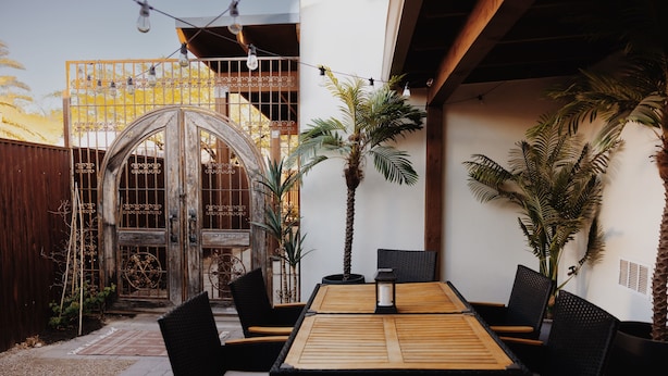 An outdoor patio with a wooden table and chairs, surrounded by potted plants and a decorative gate.