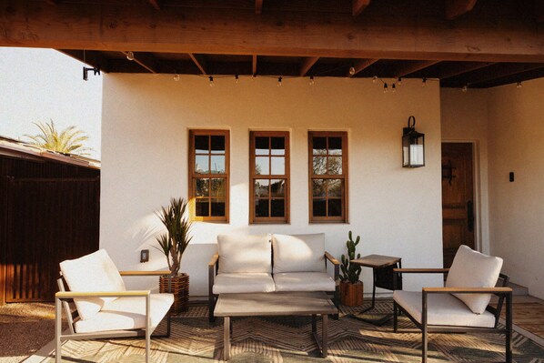 A patio with a white sofa, two armchairs, and a small table. There are three windows with wooden frames and a hanging lantern.