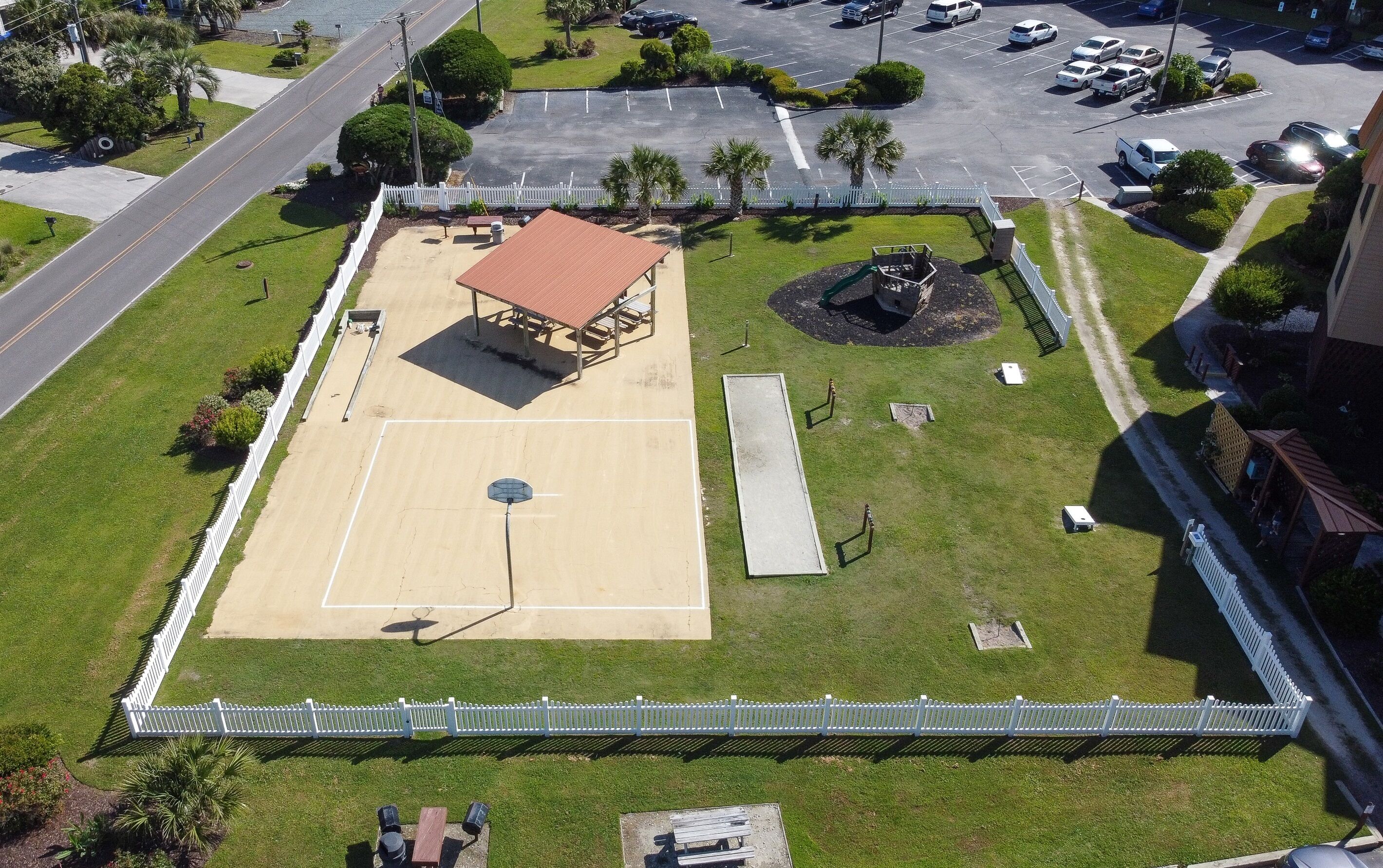 Eine Vogelperspektive zeigt einen Basketballplatz mit überdachter Zuschauertribüne, umgeben von einem weißen Zaun und Grünpflanzen.