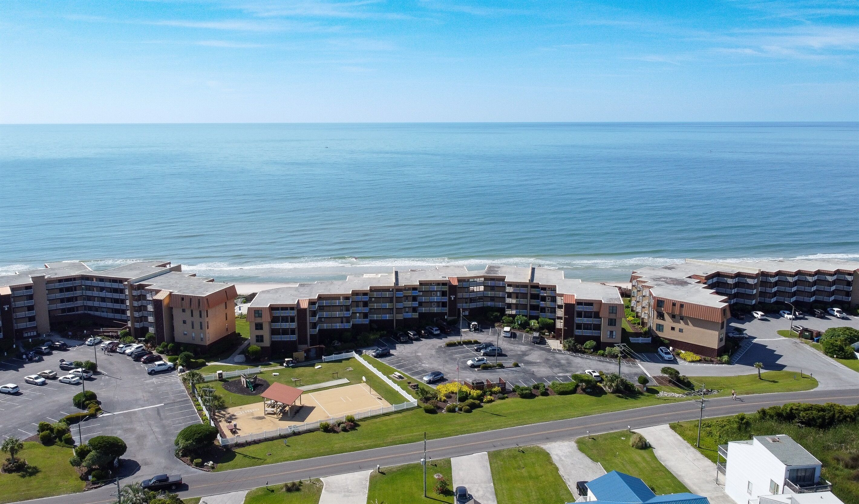 An aerial view of a coastal hotel with multiple buildings, a parking lot, and a beach in the background.