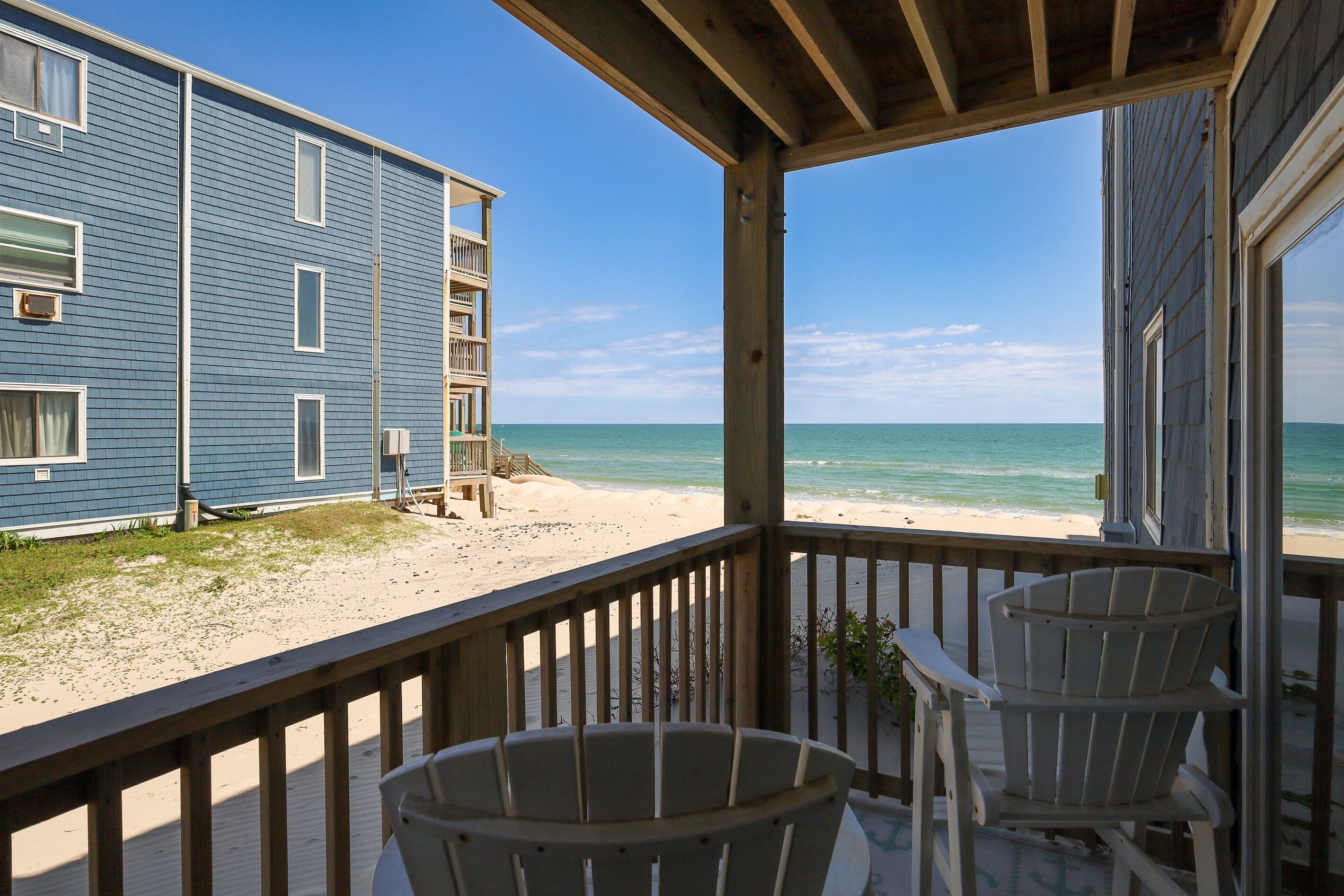Une terrasse en bois avec deux fauteuils Adirondack donnant sur une plage, avec un bâtiment visible à l’arrière-plan.