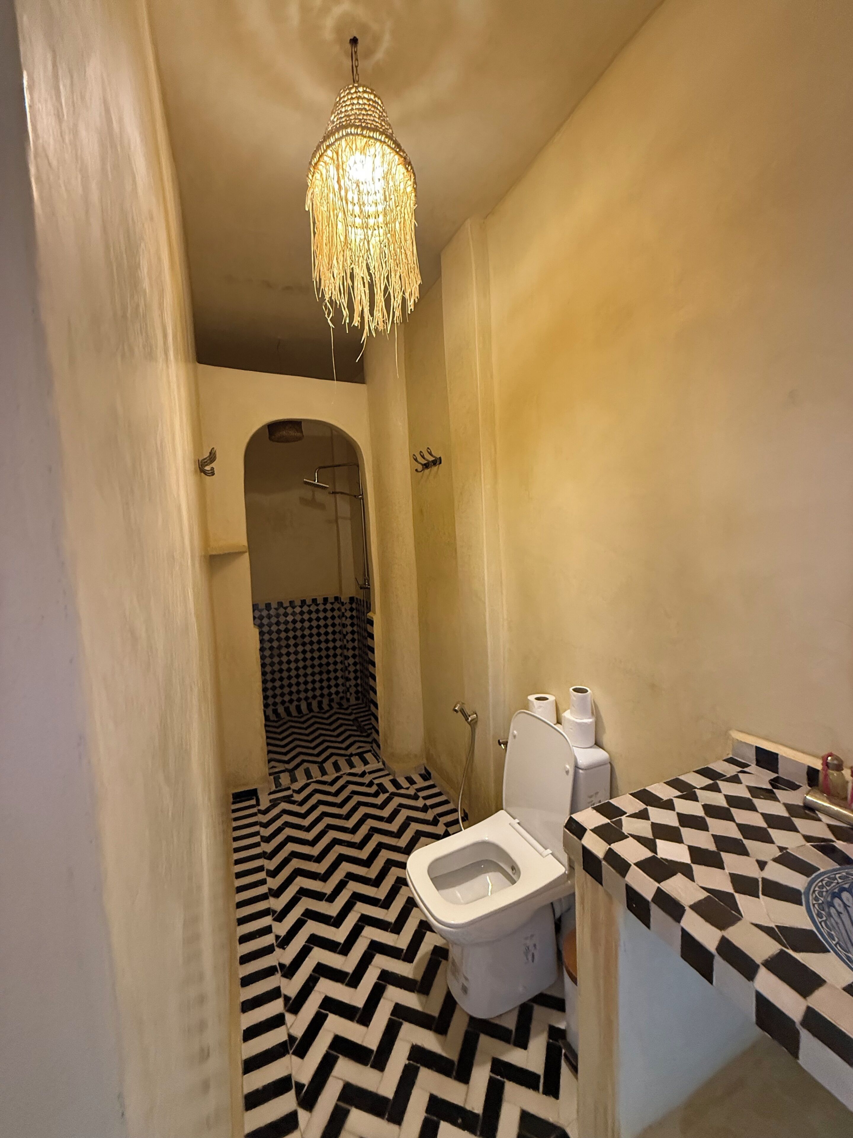 A bathroom with a black and white chevron floor pattern, a white toilet, a shower with a black and white checkered shower curtain, and a hanging light fixture.