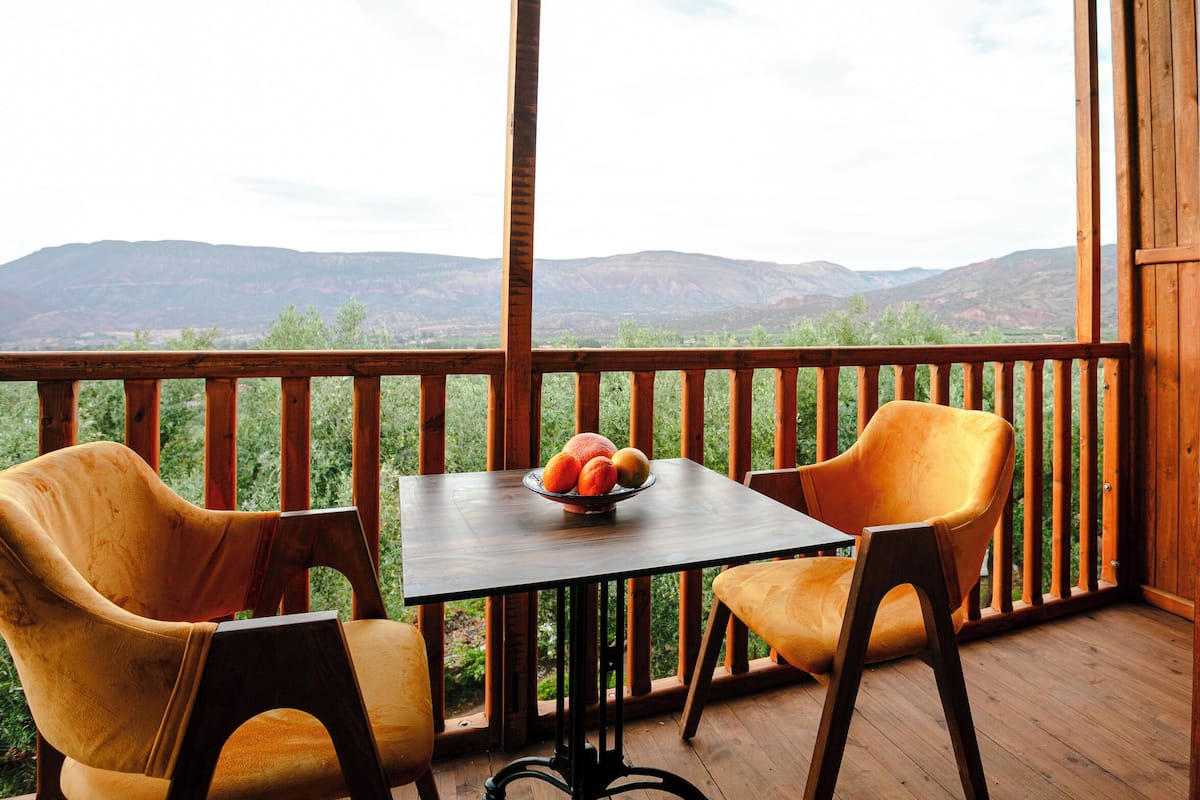 Une terrasse en bois avec deux chaises et une table, un saladier rempli de fruits et une vue sur des montagnes lointaines.
