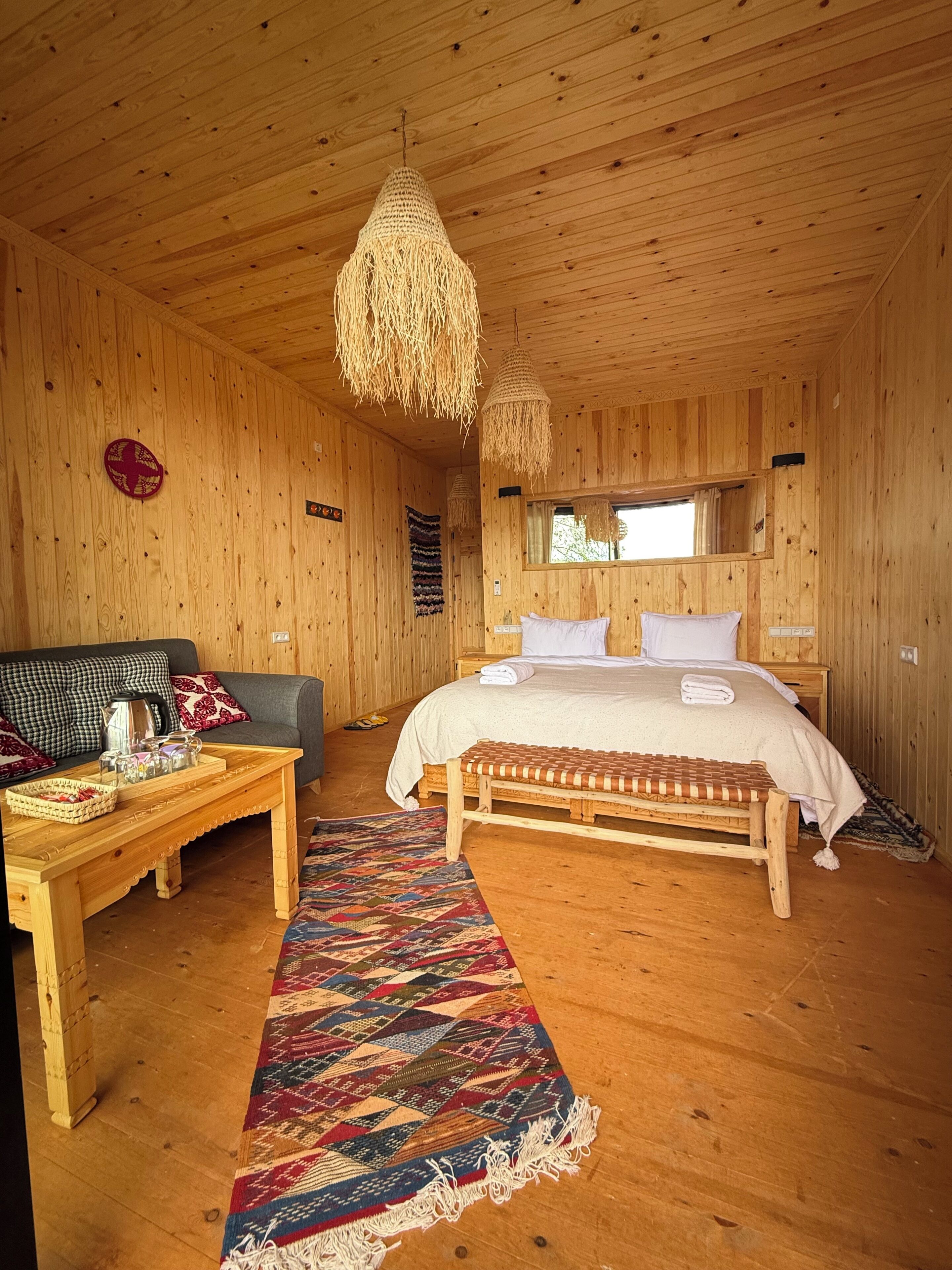 Interior de una cabaña de madera con cama, sofá, mesa y lámpara colgante.
