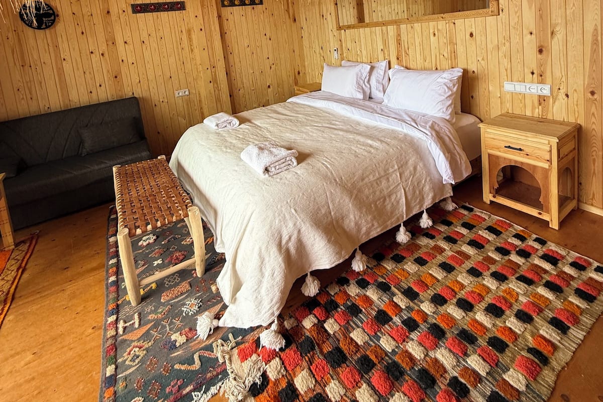 Une pièce aux murs lambrissés, comprenant un lit, un canapé, une petite table de chevet en bois et un luminaire suspendu en fibres tressées.