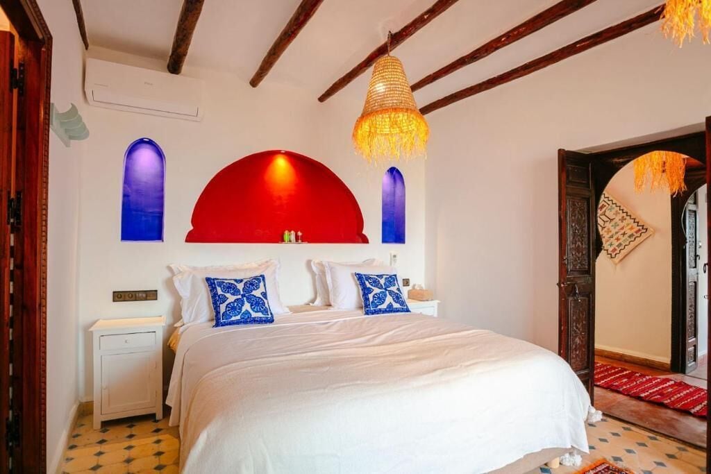 Un dormitorio con cama, mesita de noche, pared de acento roja y lámpara colgante.
