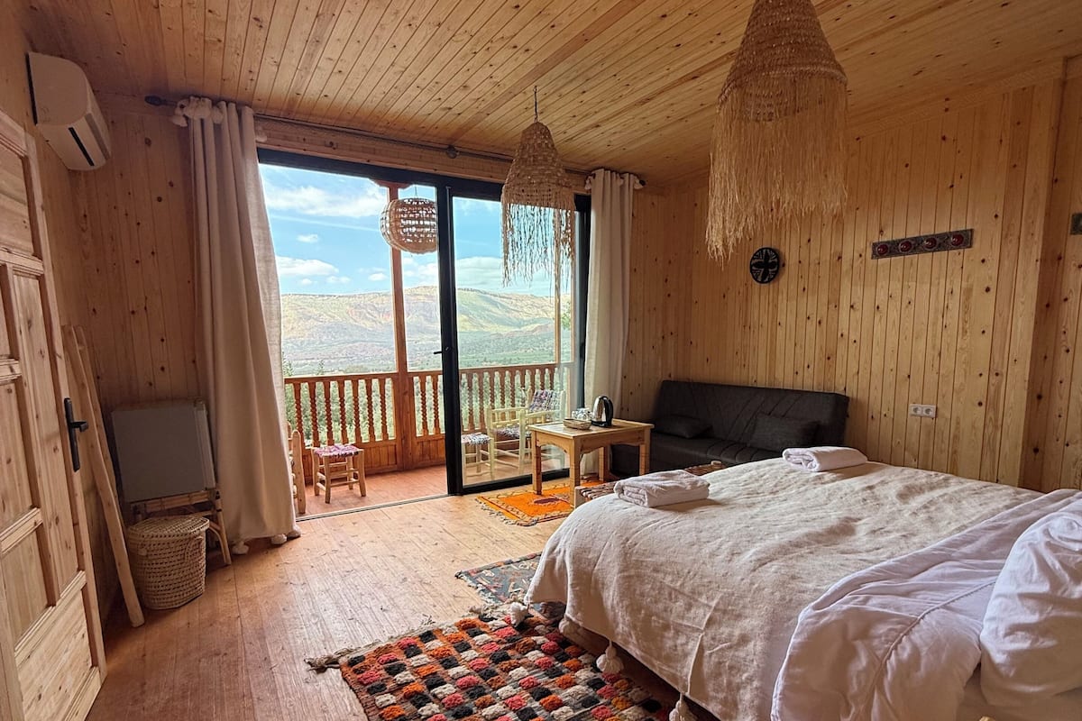 Une chambre dans une cabane en bois, avec un lit, une petite table, une chaise, un panier en osier et une vue sur les montagnes par la fenêtre.