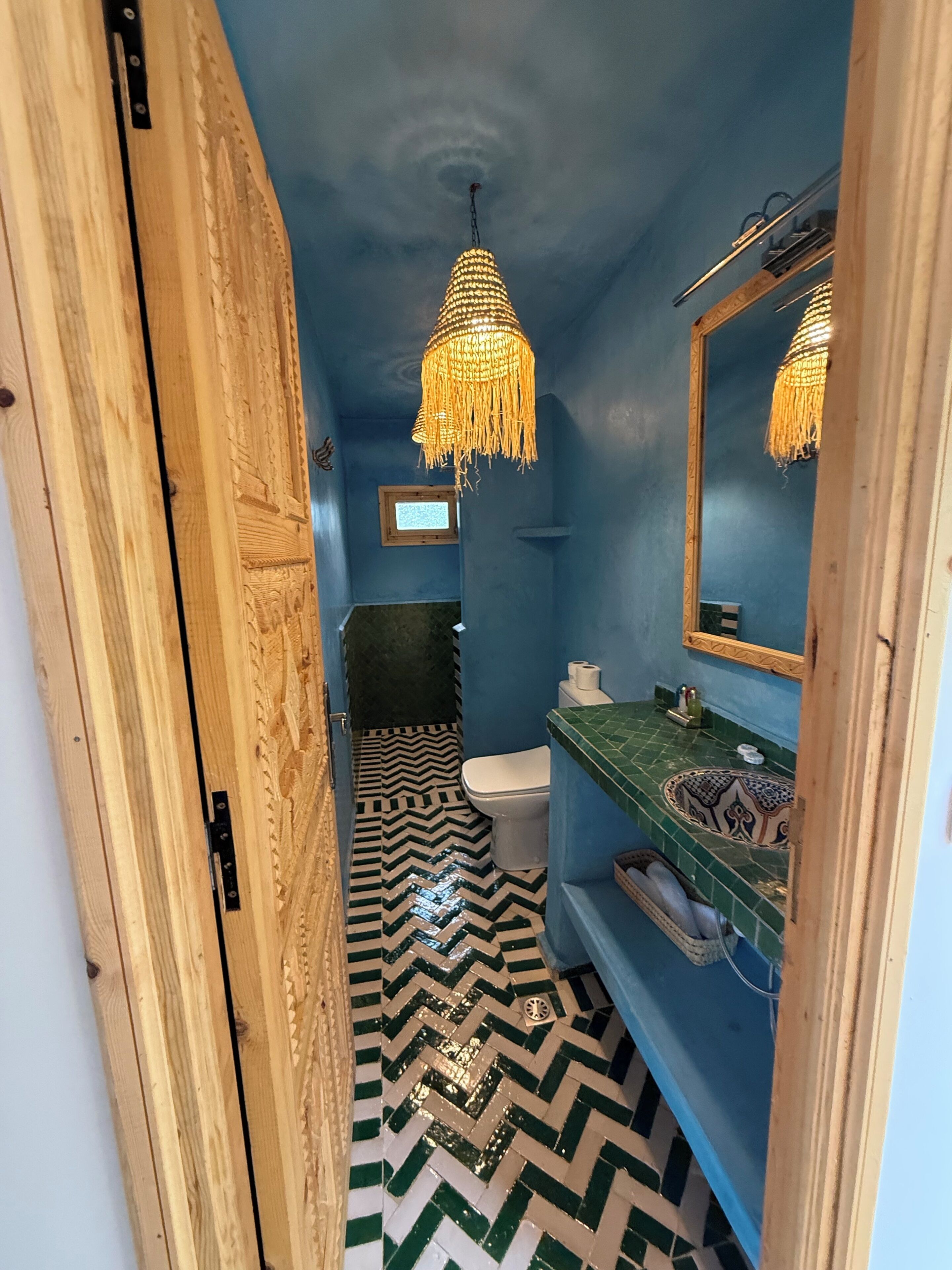 Baño con pared azul, puerta de madera, inodoro blanco, lavabo verde, piso estampado y lámpara colgante.