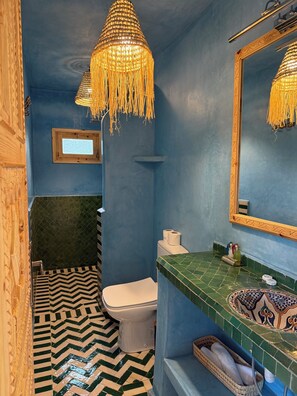 Ein Badezimmer mit blauer Wand, einer weißen Toilette, einem grün gemusterten Waschbecken und einer hängenden Leuchte.