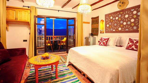 Une pièce avec un lit, un canapé rouge, une table avec des fruits, un tapis à motifs et une vue sur l’extérieur, visible à travers une porte vitrée.