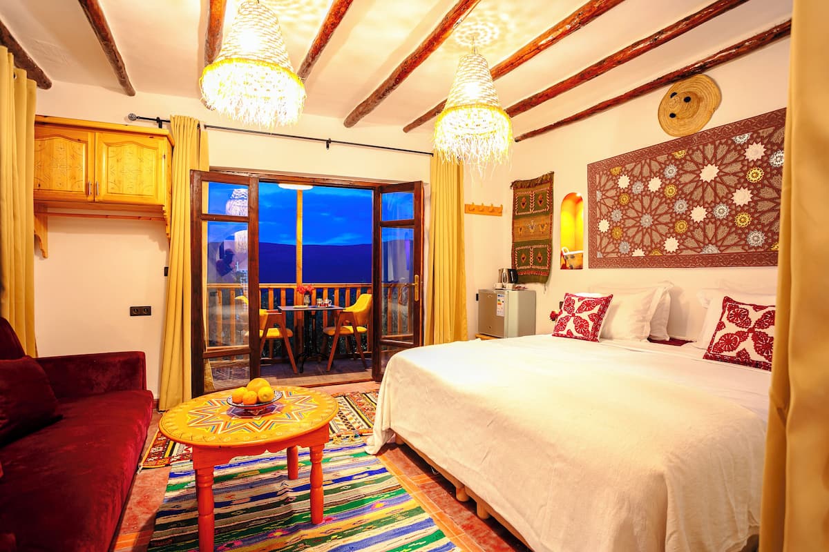 Une pièce avec un lit, un canapé rouge, une table avec des fruits, un tapis à motifs et une vue sur l’extérieur, visible à travers une porte vitrée.