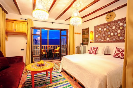 A room with a bed, a red sofa, a table with fruit, a patterned rug, and a view through a glass door.