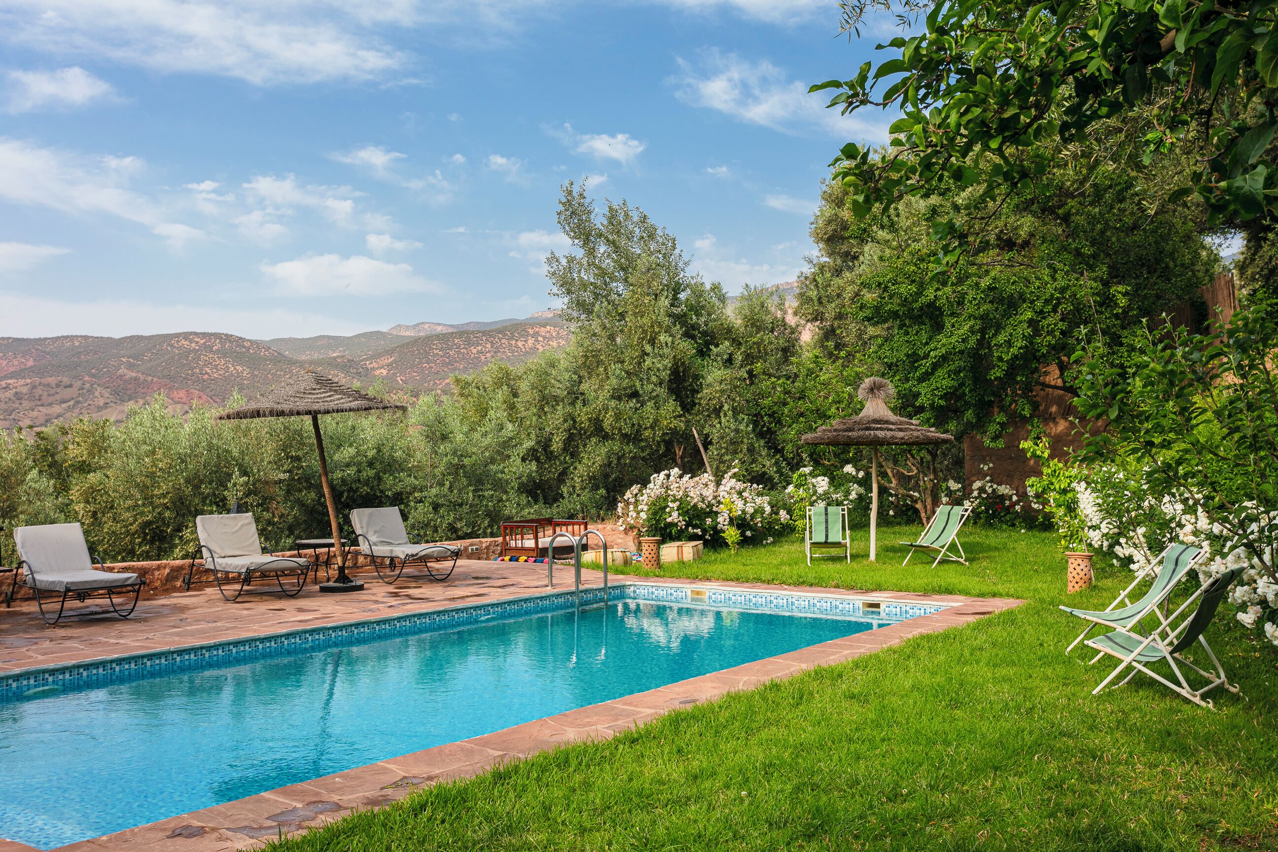Uma piscina com espreguiçadeiras e um pérgola, cercada por vegetação exuberante e colinas ao fundo.