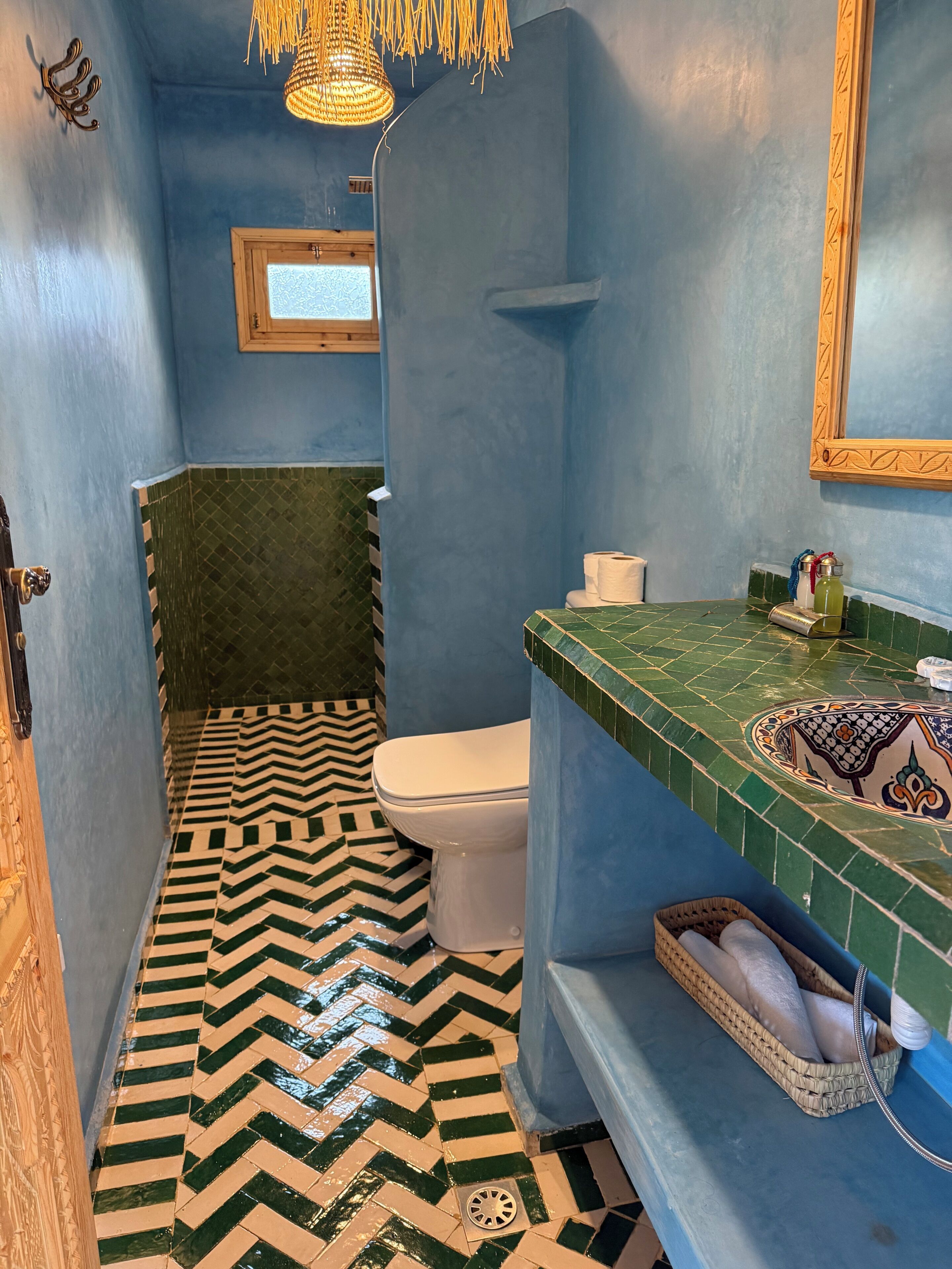 Un baño con pared azul, inodoro blanco, suelo con estampado verde y blanco, lámpara colgante y zona de lavamanos con azulejos.
