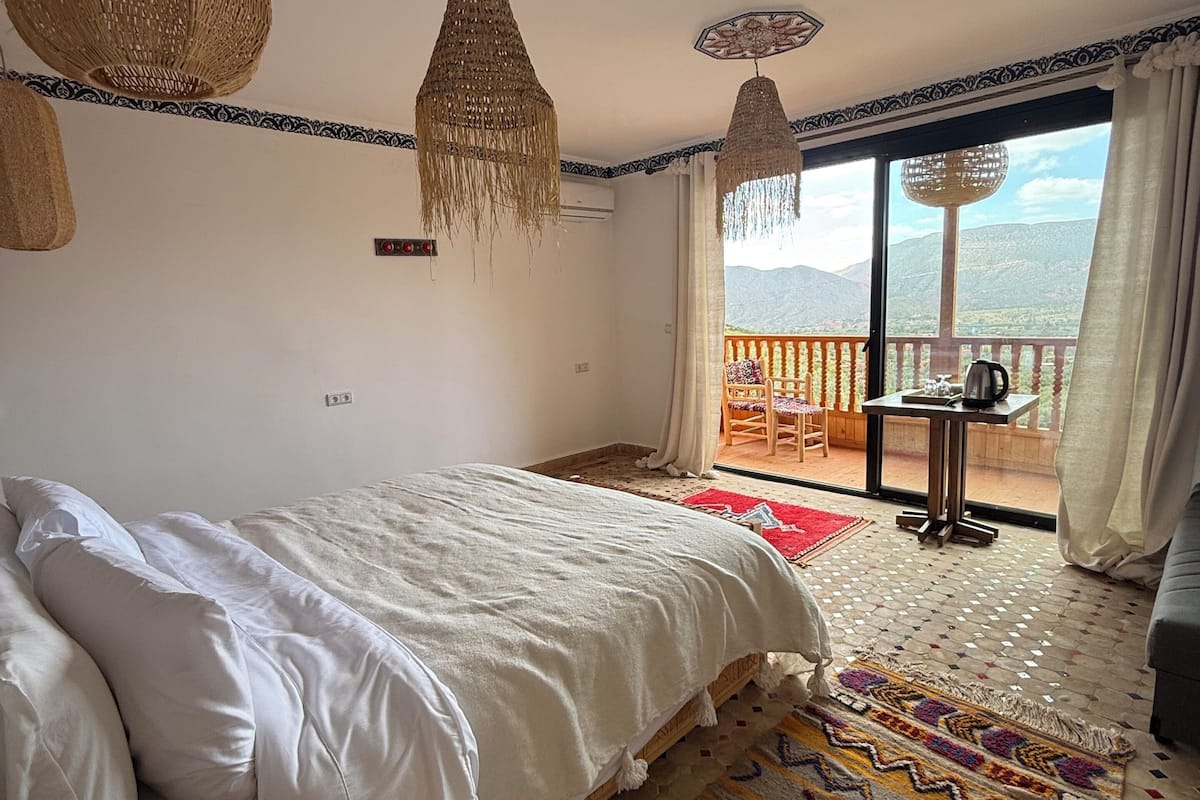 Une chambre avec un lit, une table de chevet en bois, un tapis à motifs, un balcon avec une table et des chaises, et une vue sur les montagnes.