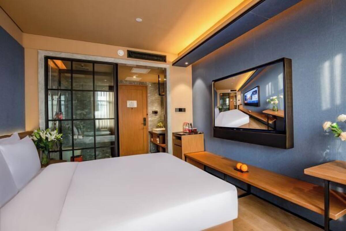A modern hotel room with a bed, a TV mounted on the wall, a wooden bench, and a small table.