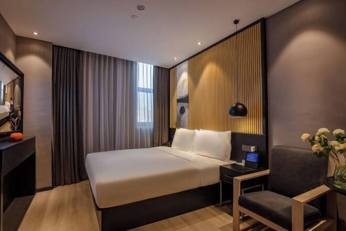 A modern hotel room with a large bed, a desk with a computer, a chair, and a bedside table.