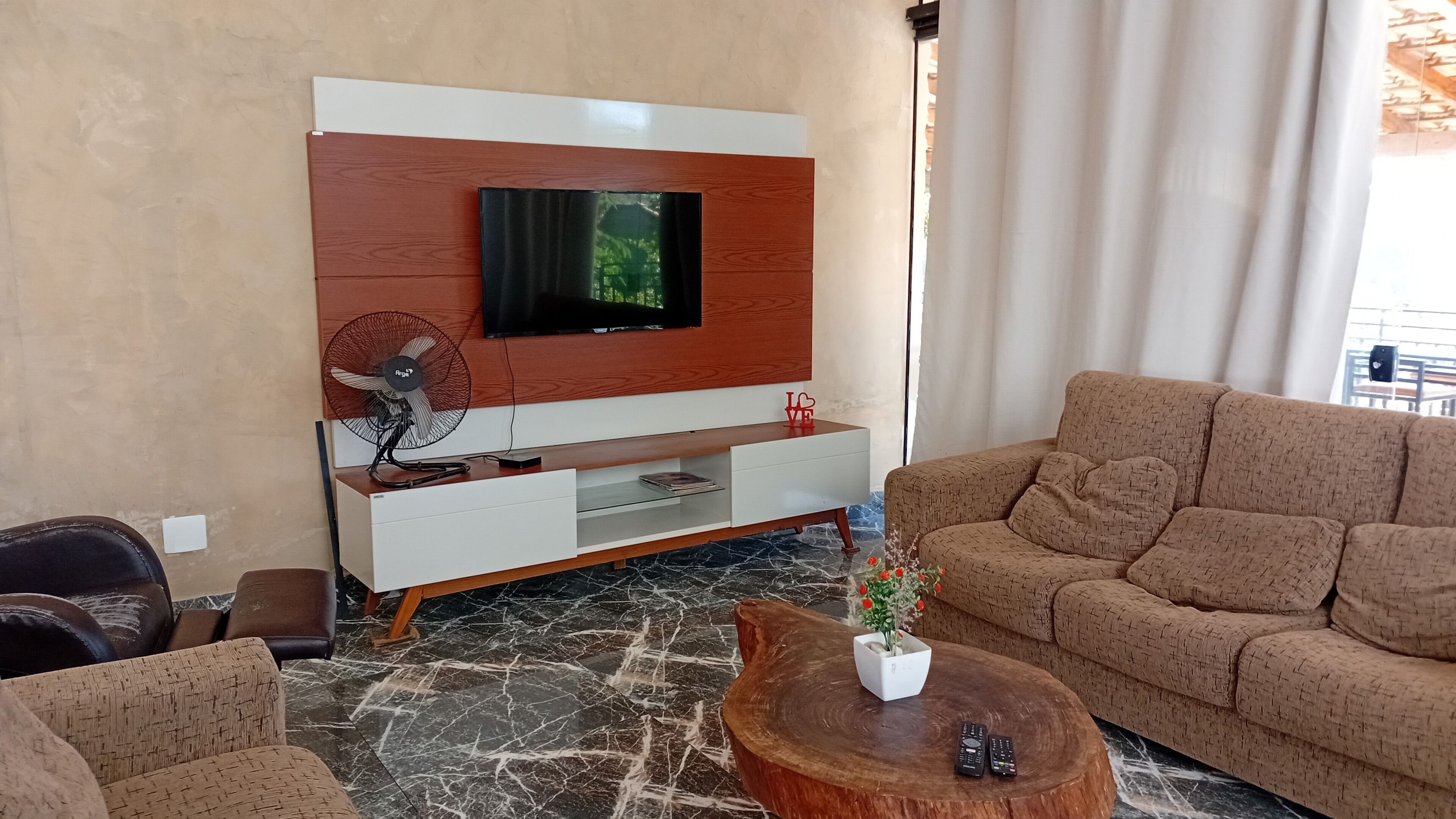 Ein modernes Wohnzimmer mit einem flachbildfernseher an einer Holzwand, einer weißen Unterhaltungsanlage, einem braunen Sofa, einem Couchtisch mit einer Pflanze und einem Ventilator.