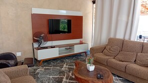 A modern living room with a flat-screen TV mounted on a wooden wall, a white entertainment unit, a brown sofa, a coffee table with a plant, and a fan. - Country house/villa with a pool, sports field, and hot tub; pets allowed. Best price in the region. (Itu)