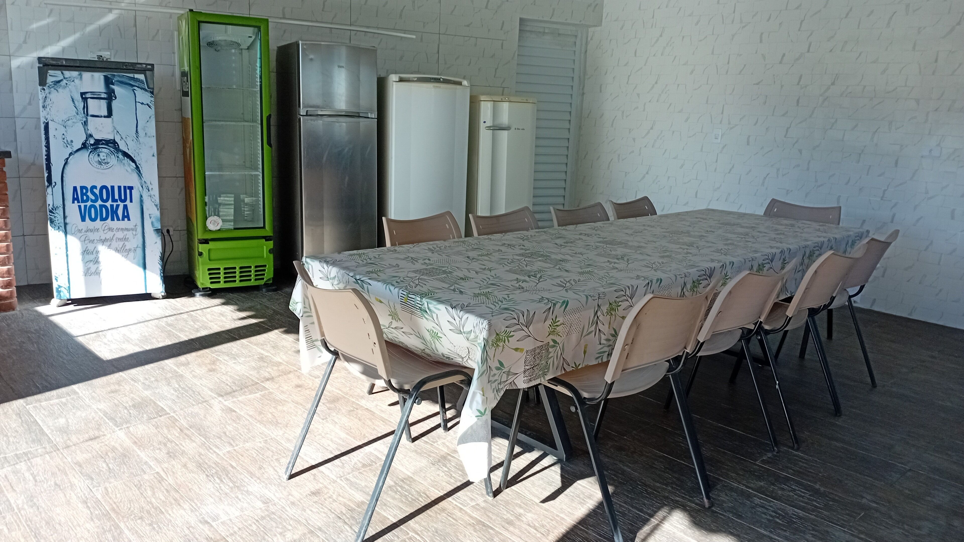 A dining area with a table set for six, surrounded by chairs. There are two refrigerators and a green appliance, possibly a cooler, in the background.