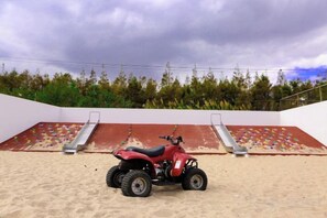 Un vehículo todo terreno rojo sobre una superficie arenosa, con una pared de escalada y una escalera de fondo.