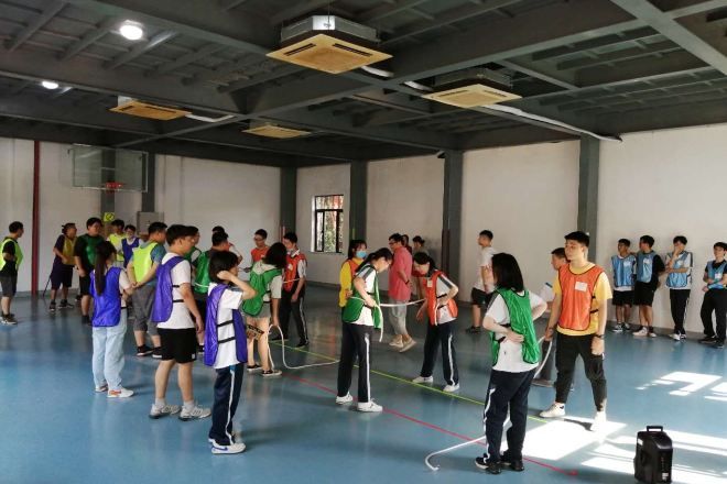 Un grupo de personas en un gimnasio, con un aro de baloncesto, forman una fila, posiblemente esperando un evento.
