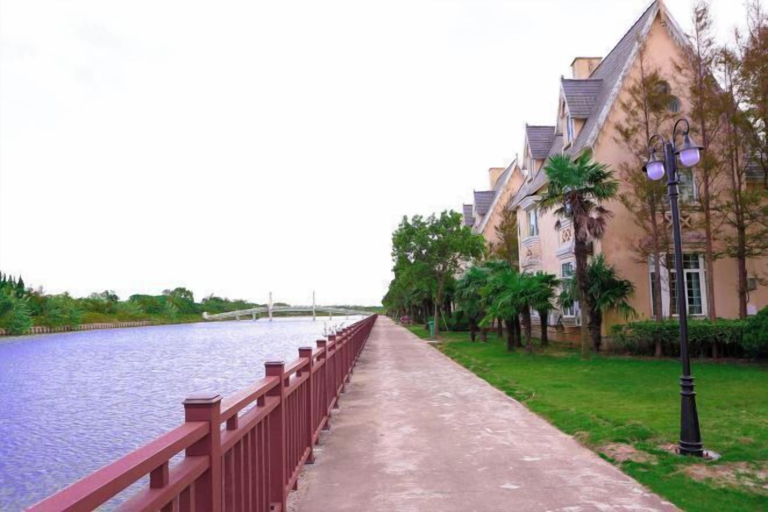 A riverside walkway with a red railing, lined with trees and a building with a sloped roof.