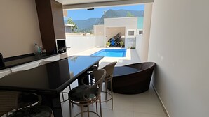 A modern kitchen with a black countertop, two wooden chairs, and a view of a swimming pool and mountains in the background. - Casa com piscina em Boraceia (Bertioga)