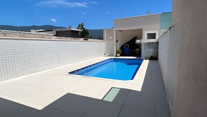 A modern outdoor swimming pool with a tiled deck, surrounded by a low white wall and a concrete structure. - Casa com piscina em Boraceia (Bertioga)