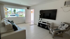 A modern living room with a white sofa, a patterned armchair, a flat-screen TV mounted on the wall, and a sliding glass door leading to a balcony. - Casa com piscina em Boraceia (Bertioga)